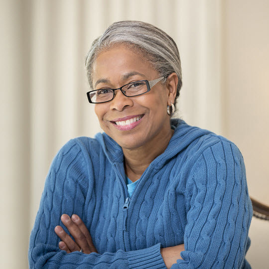 An older woman wearing glasses and a blue sweater, smiling gently while seated in a cozy environment.