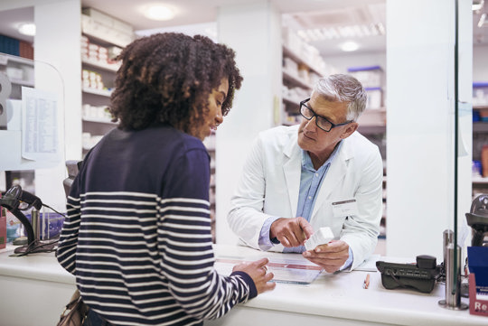 Inside a pharmacy, a man and woman discuss items while standing near shelves filled with various pharmaceutical products.