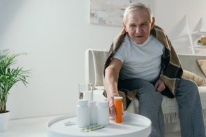 An older man sits on a couch, surrounded by pills and a cozy blanket, reflecting a moment of rest and contemplation.