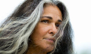 An older woman with long hair wearing a gray shirt, smiling gently while standing in a serene outdoor setting.