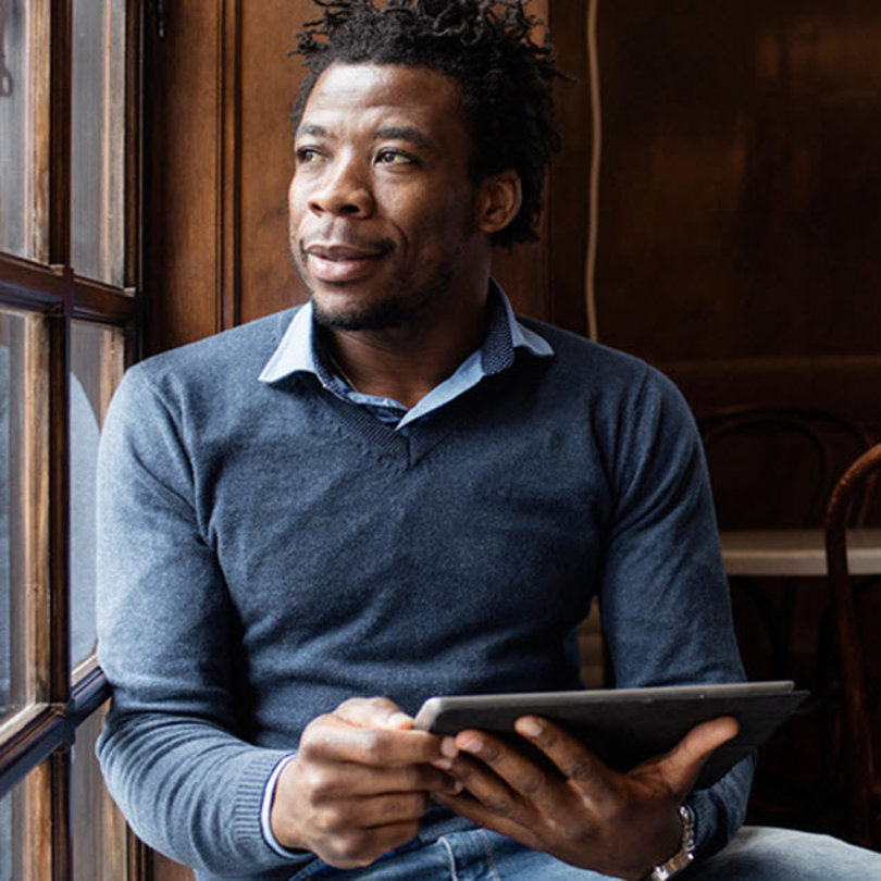  A man wearing a sweater and jeans is seated at a table, engaged with a tablet.
