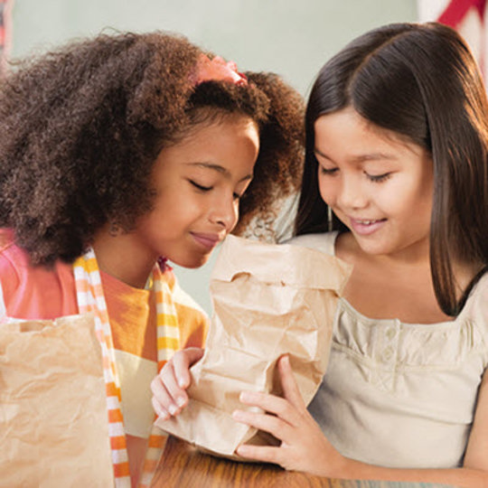 Two young girls joyfully holding brown paper bags, smiling as they stand together outdoors.